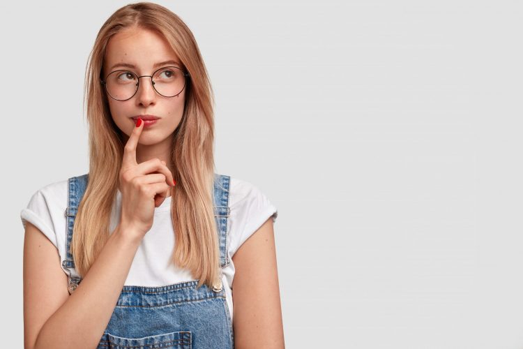 Pretty blonde female with focused expression makes choice in mind, recalls some moment in life, keeps fore finger near mouth, stands against white wall with copy space for your advertisement