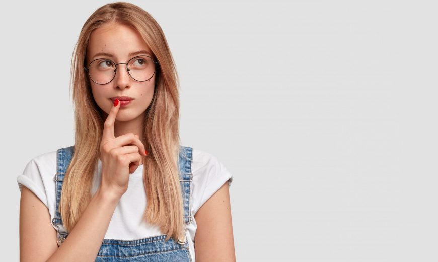 Pretty blonde female with focused expression makes choice in mind, recalls some moment in life, keeps fore finger near mouth, stands against white wall with copy space for your advertisement